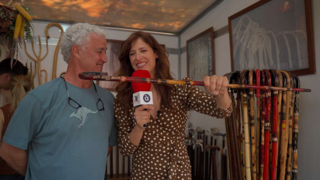 Reporter interviews a smiling man in a blue t-shirt as she holds a red microphone, showing a long decorative staff between them, with canes on display in the background.