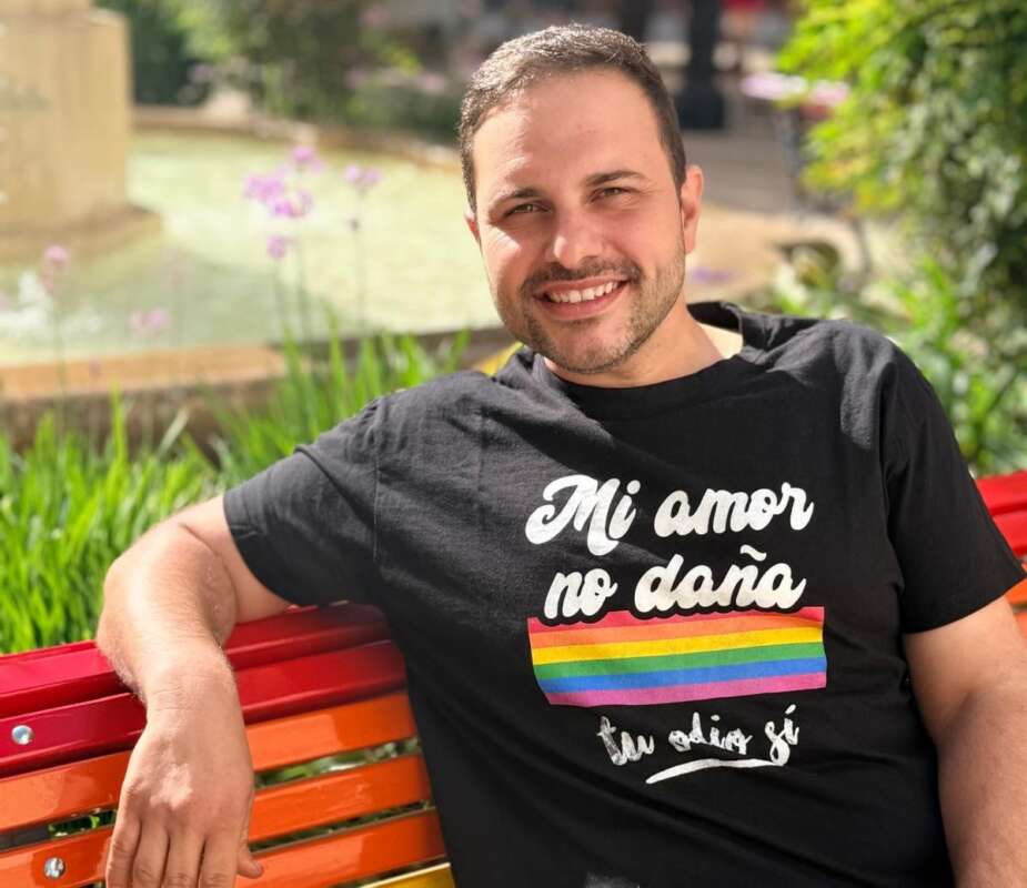Man smiling while sitting on a red bench outdoors, wearing a black shirt with a rainbow flag and Spanish slogan.