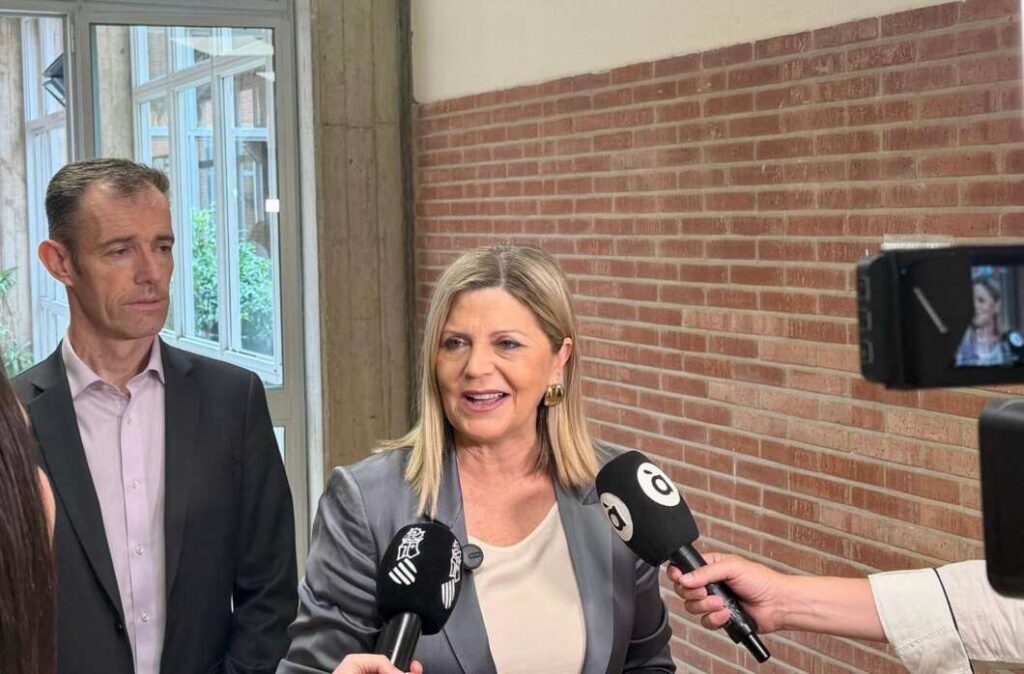 Woman in a gray blazer speaks to reporters, several microphones in front, a man in a suit stands beside her near a brick wall.
