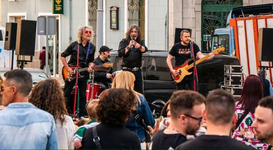 A four-piece street band performing on a sidewalk stage with guitars and drums while a crowd watches nearby.