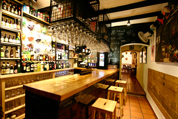 Cozy bar interior with a long wooden counter, hanging glassware, bottles on illuminated shelves, and a chalkboard wall at the back.