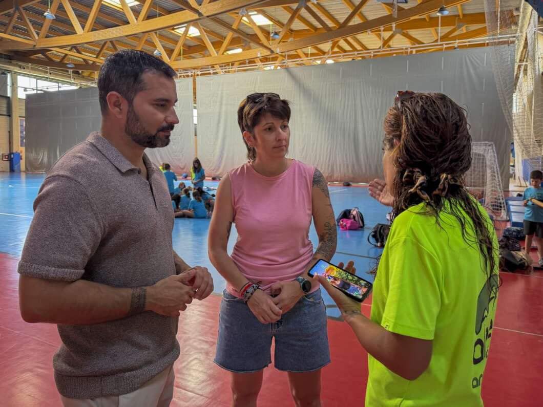 Three adults in a gymnasium stand chatting: man in a polo, woman in a pink sleeveless top, and a person in a neon shirt showing a phone to them, with kids sitting in the background.