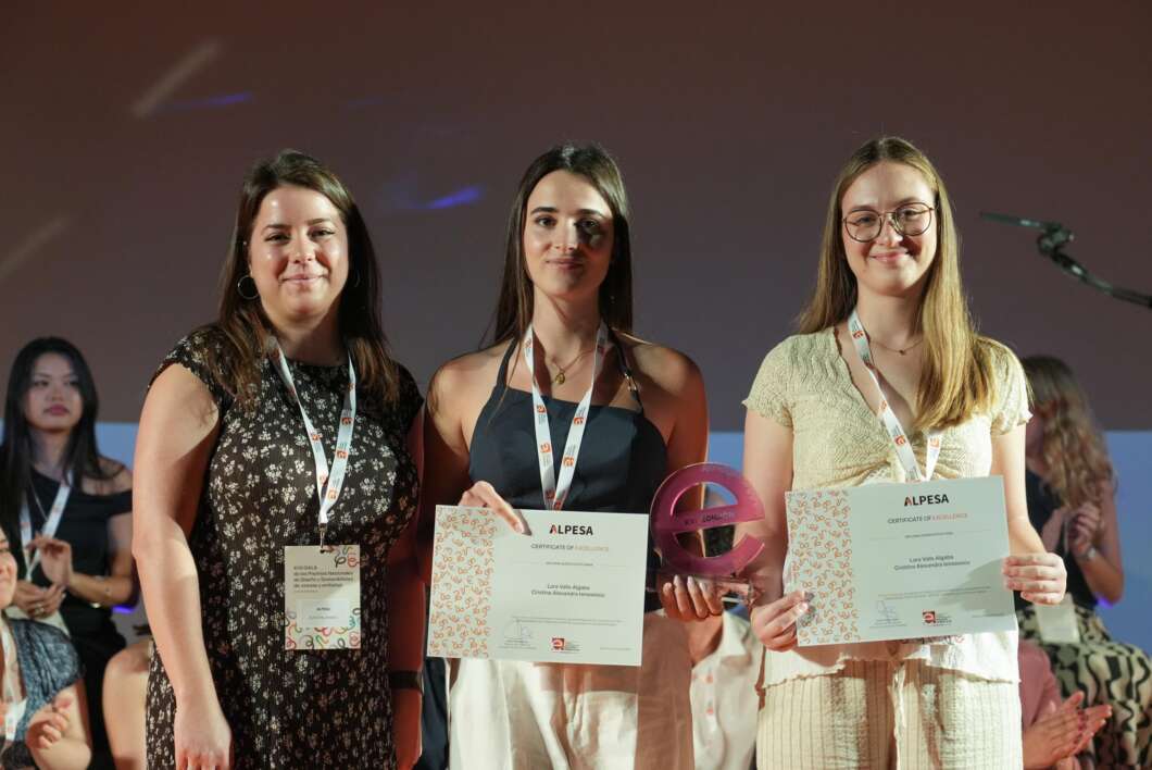 Three young women on a stage holding certificates and a pink trophy, smiling at the camera with lanyards around their necks, awards event.