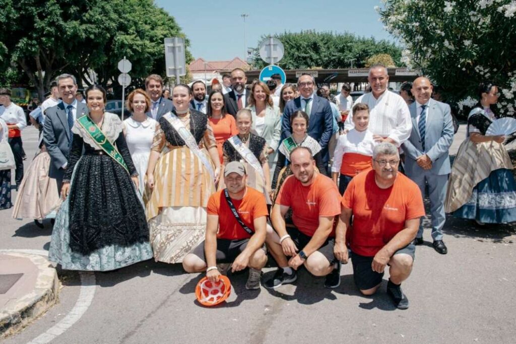 Group of people posing on a street, some in traditional dresses and others in orange volunteer shirts, likely at a cultural festival or parade.