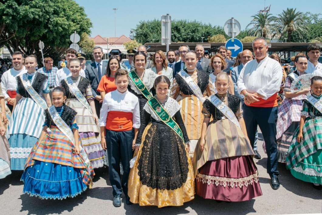 Group of people in colorful traditional dresses posing at a sunny street festival or parade, with sashes and ornate gowns in the foreground.