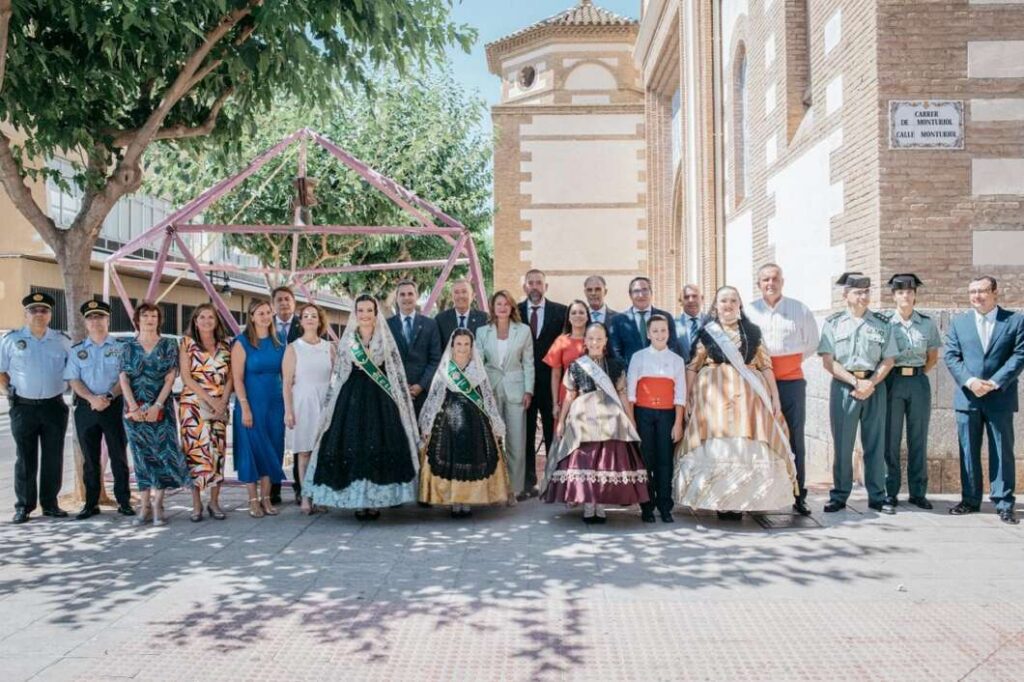 Group of men and women in formal attire, some wearing sashes, posing for a sunny outdoor group photo in a plaza with a brick building behind.
