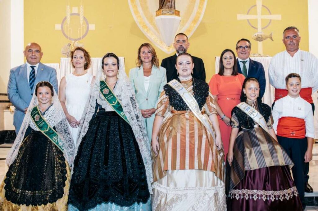 Group of people in formal attire and traditional gowns posing indoors, likely at a ceremony or pageant with a yellow wall and religious symbols behind them.