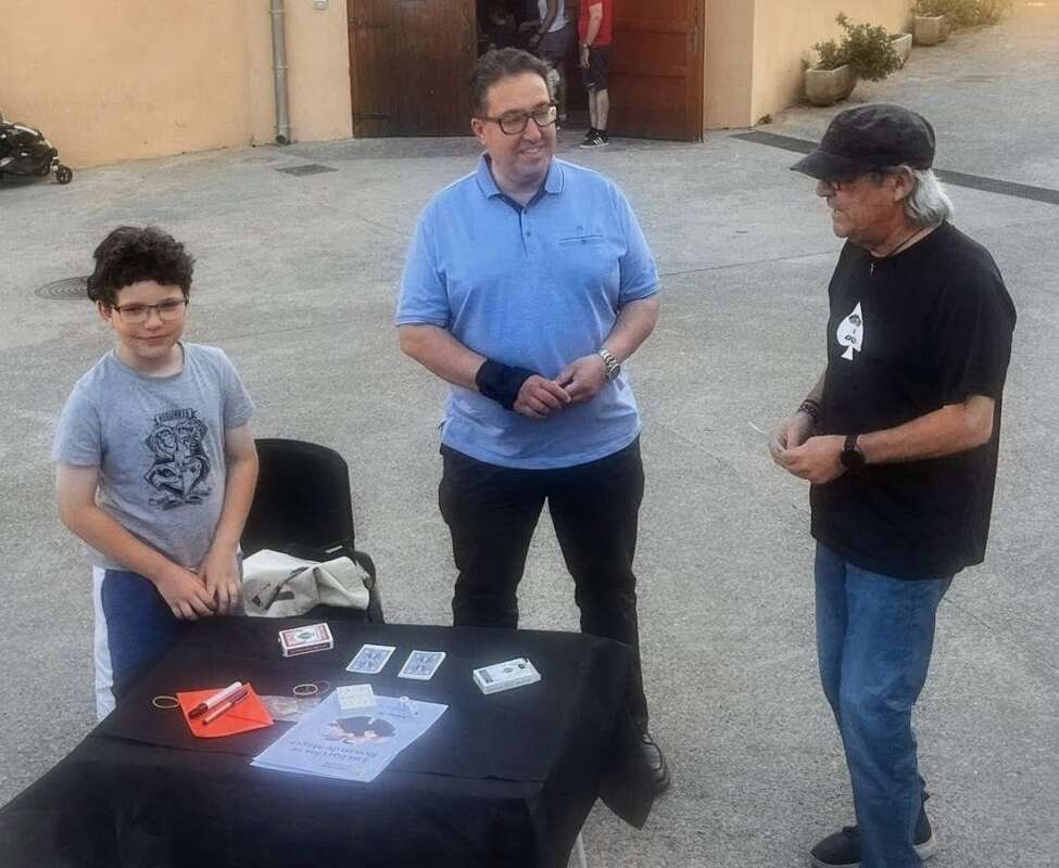 Three people stand around a black table with playing cards and a flyer, outdoors near a beige building.