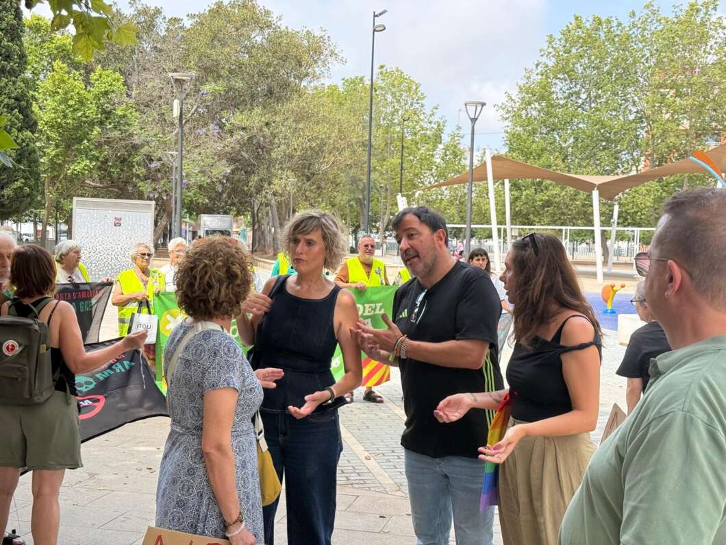 Group of people chatting in a park at a community event, with volunteers in yellow vests behind a barrier.