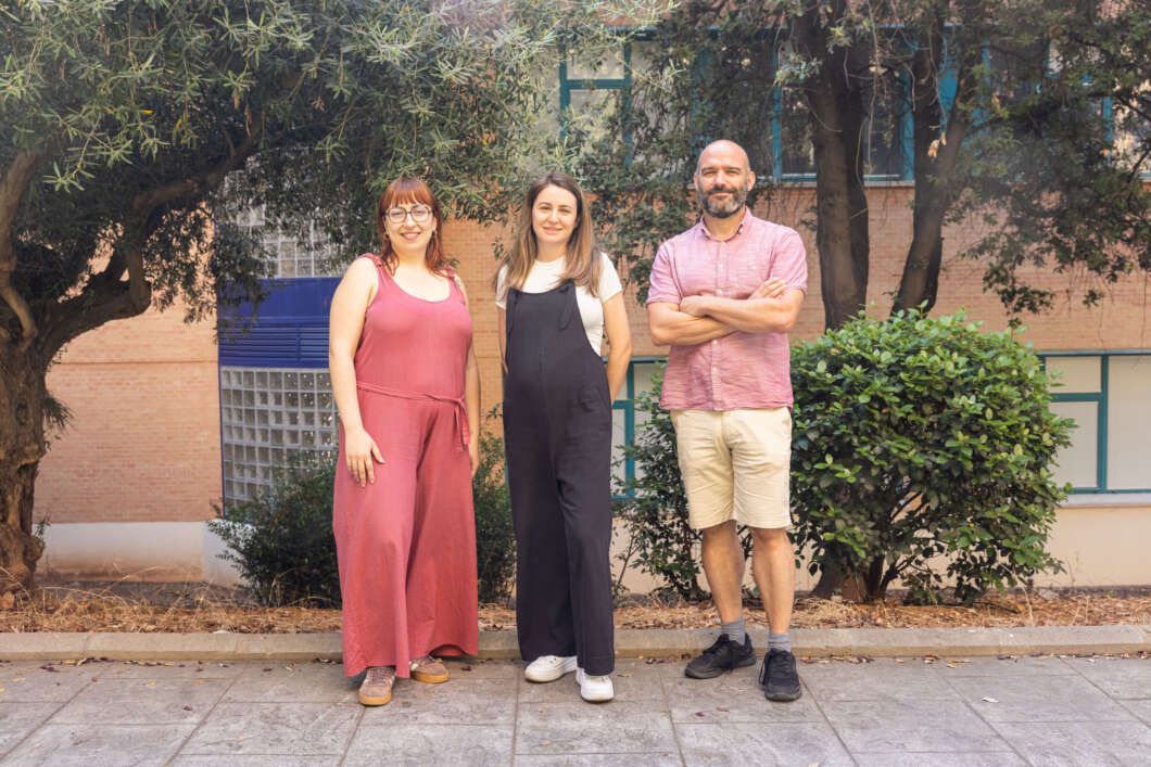 Three people stand together outdoors in front of a brick building with trees, smiling for the photo.
