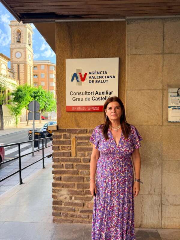 Woman in a purple patterned dress standing outside a health agency sign (Agència Valenciana de Salut) on a sunny street.