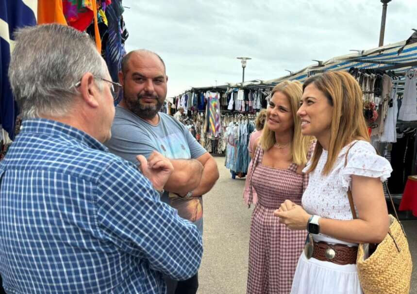 Group of four adults talking at an outdoor clothes market; two women smile while a man in a gray t-shirt stands with folded arms near market stalls.