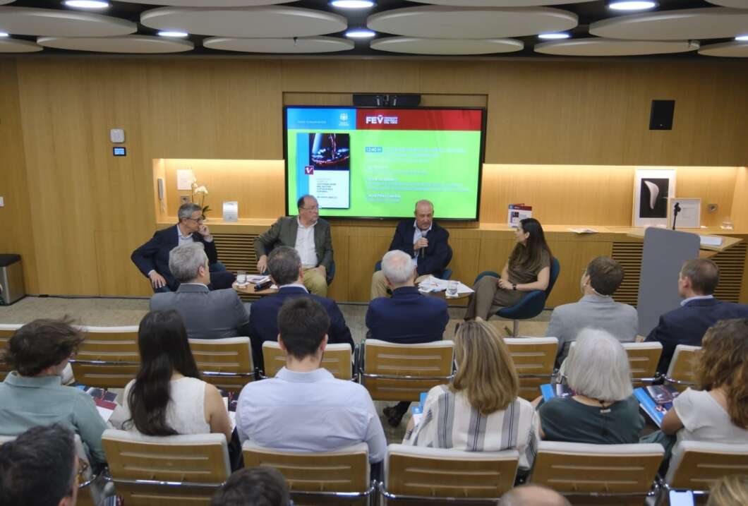 Panel discussion in a lecture room with five people seated on a stage and a large screen behind them displaying a green slide; audience seated in rows faces the panel.