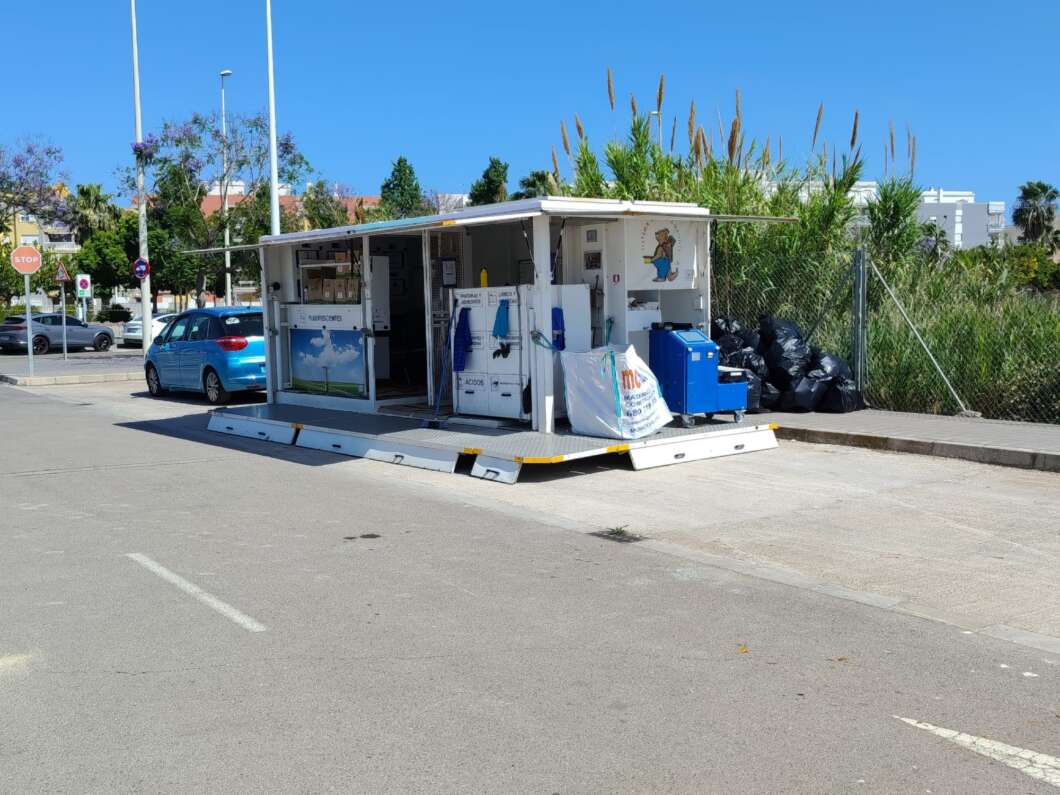Mobile recycling trailer in a parking lot, with bins and bags of trash nearby under a clear sky.