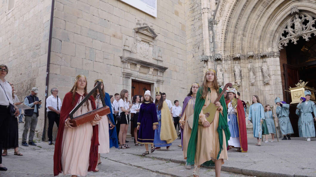 Group of people in medieval costumes outside a stone church, with a young woman in a green cape carrying a lute at the front