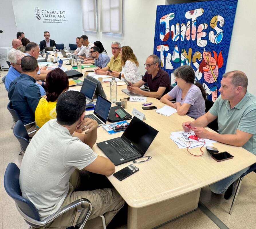 Group of people around a long conference table with laptops and papers in a bright room; Generalitat Valenciana banner in the background.
