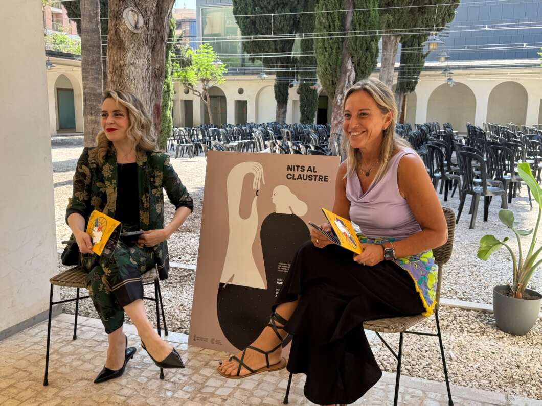 Two women seated outdoors at a book event, each holding a yellow book; a large poster between them reads 'NITS AL CLAUSTRE'.