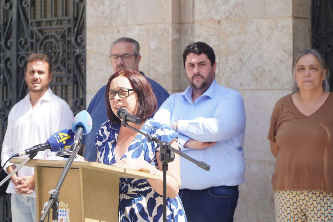 Woman speaking at a podium with microphones, addressing a press conference outside a stone building, with four people standing behind her.