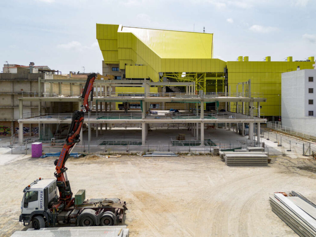 Construction site with a multi-level concrete structure in progress and a large yellow building in the background; a red crane is on site.
