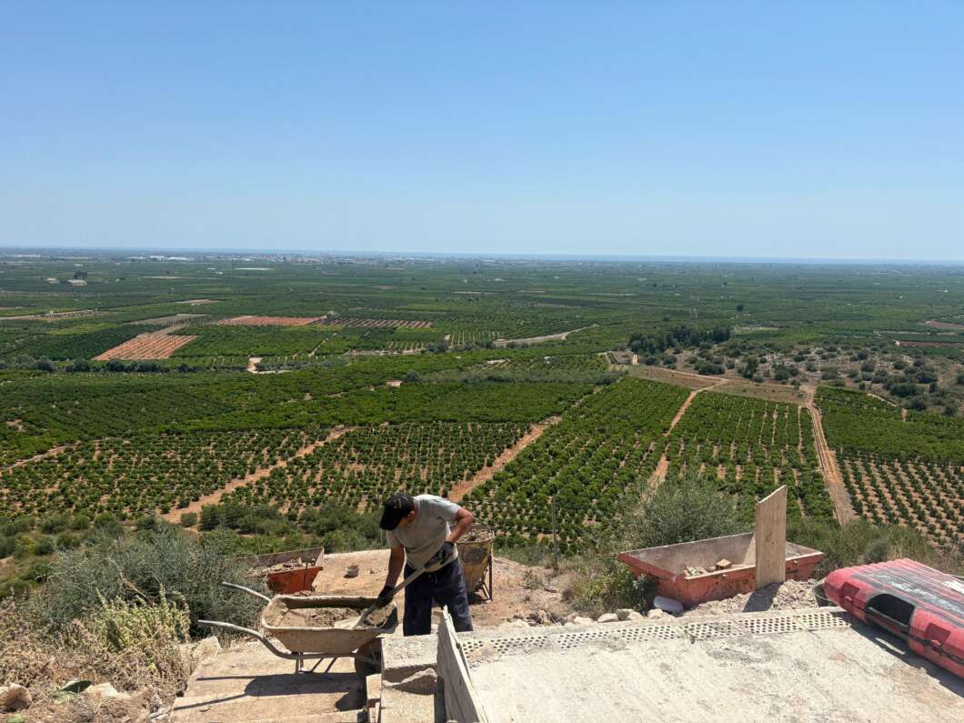 Worker pushing a wheelbarrow on a construction site at the edge of a hill, overlooking expansive farm fields and orchards under a clear blue sky.