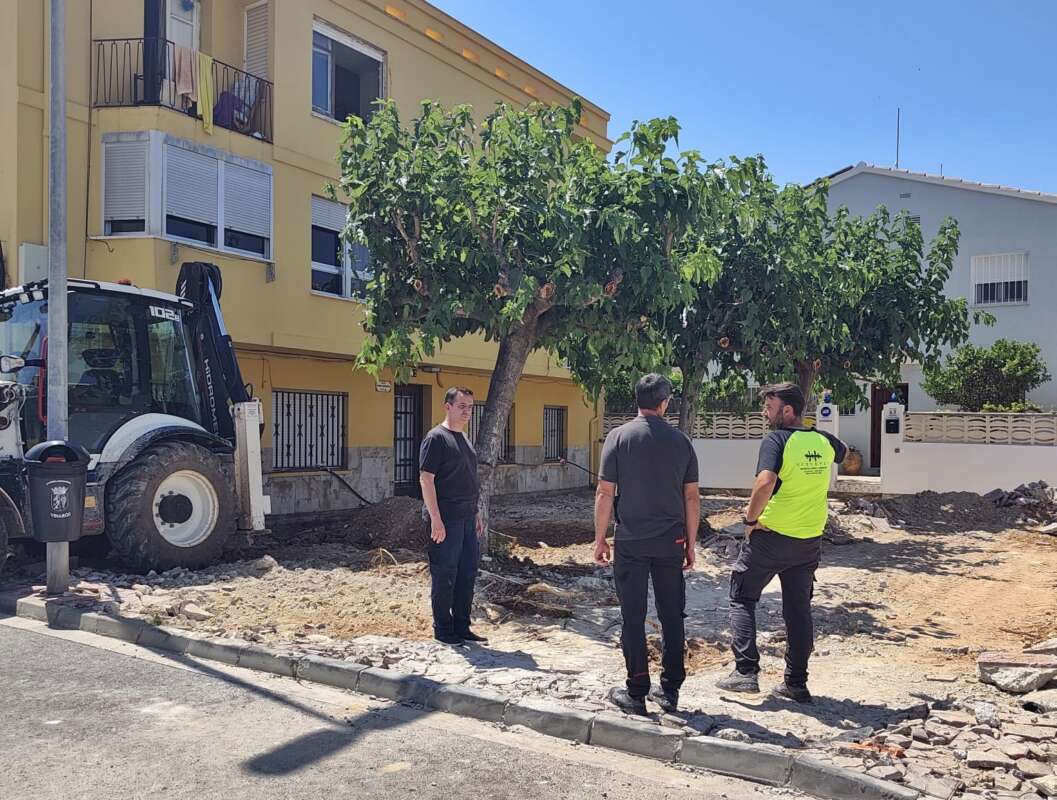 Three men stand on a damaged curb and rocky ground beside a tree and a parked construction vehicle, in a residential area with yellow buildings.