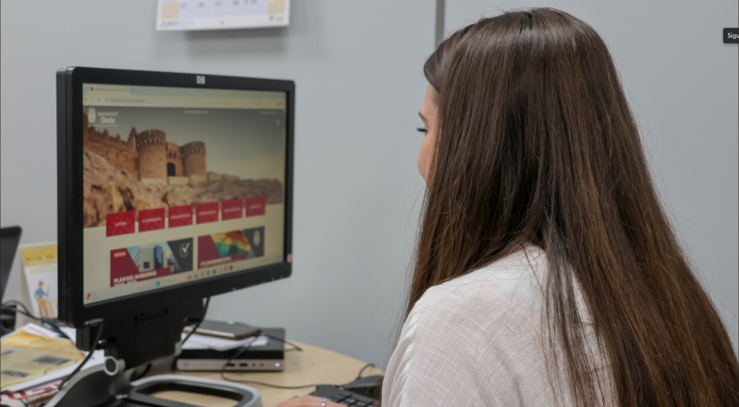 Woman with long brown hair sits at a desk, looking at a computer monitor displaying a website with a fortress image on the home page.
