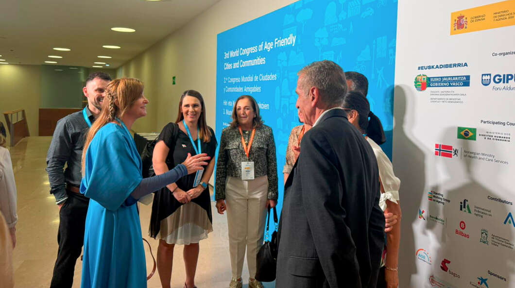 Group of professionals wearing lanyards chat in a hallway beside a blue conference backdrop with event title.