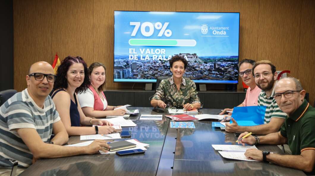 Eight people sit around a conference table in a meeting room, with a large screen showing '70%' and 'El valor de la palma' in the background.