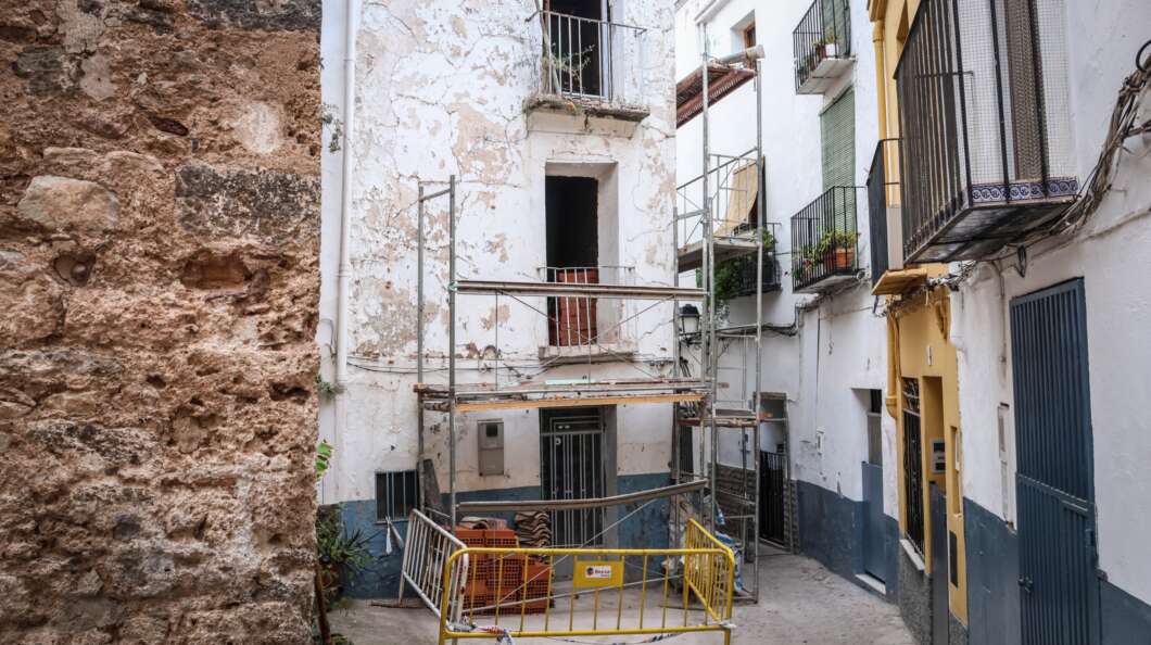 Narrow street with old white building under renovation; metal scaffolding blocks entry and yellow barriers surround a small construction area.