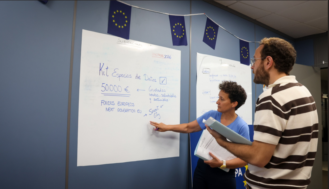 Two people discuss a whiteboard filled with notes in a room decorated with European Union flags above.
