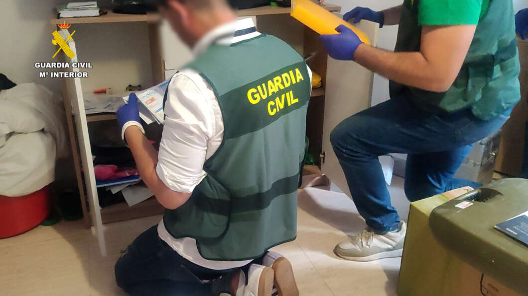 Guardia Civil agents in a cluttered room, one kneeling and sorting papers while the other holds a yellow evidence envelope.