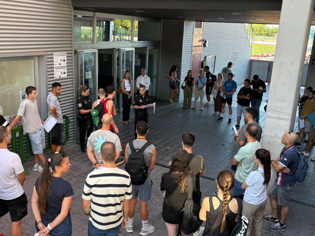 Group of people in a circle outside a building listening to a uniformed officer who is holding papers for a briefing.
