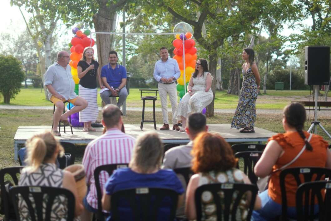Group of six people on an outdoor stage speaking to an audience at a park event with rainbow balloon decorations.