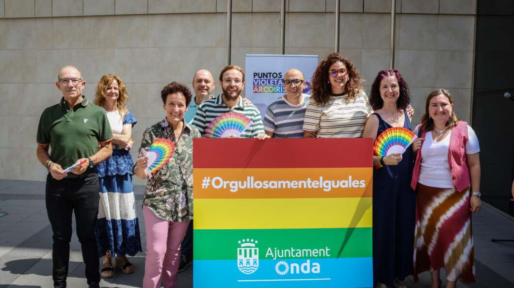 Group of smiling adults posing behind a rainbow banner that reads '#Orgullosamenteliguales' at an LGBTQ+ pride event, with 'Ajuntament Onda' logo visible.