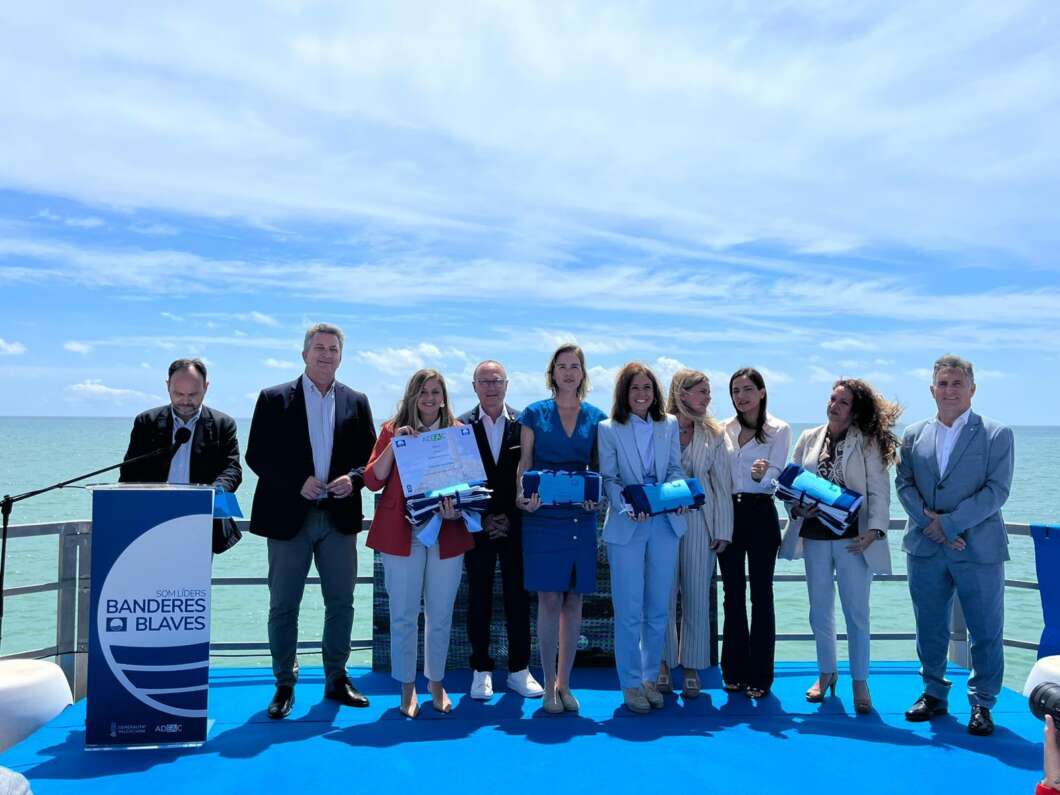 Group of ten people on a blue stage by the sea, holding certificates and blue folders during an outdoor award ceremony by the water.