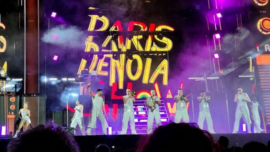 Group of performers on a brightly lit stage performing a concert, wearing white outfits with a neon signs backdrop and purple smoke effects, audience in foreground.