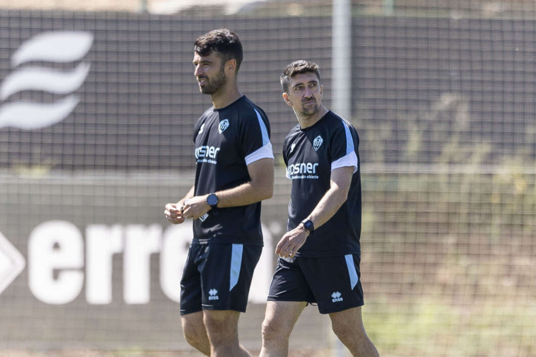 Two male soccer players in dark training kits walk on a practice field during a training session.