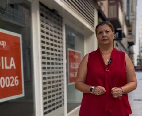Woman in a red sleeveless blouse and wide-leg black pants stands on a city sidewalk in front of a storefront with orange sale signs, looking at the camera.
