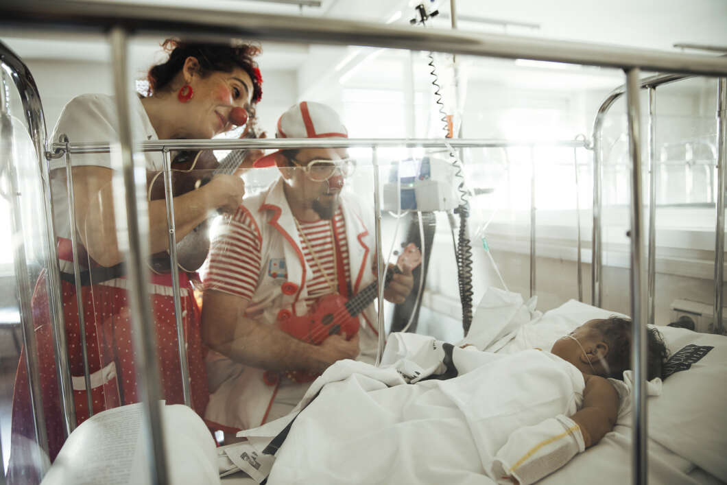 Two clowns in colorful costumes visit a child in a hospital bed, playing a red guitar beside the patient's bed.