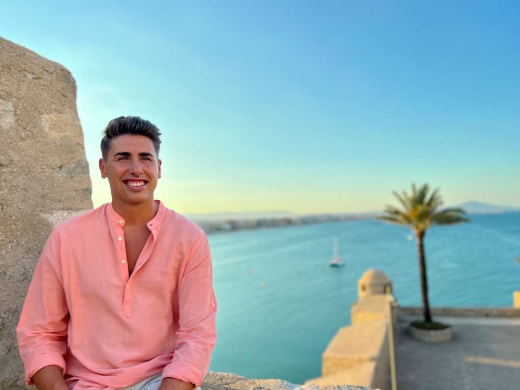 Smiling man in a pink button-down shirt sits beside a stone wall with a seaside promenade and palm tree in the background.