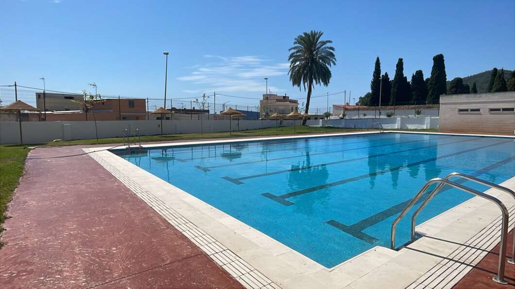 Outdoor public swimming pool with blue water, lane markings, a metal ladder, and a red deck with palm trees nearby.