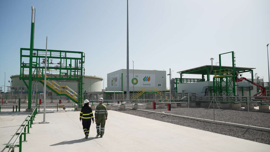 Two workers in hard hats walk along a concrete path at an industrial plant with green metal structures and pipelines on a sunny day.