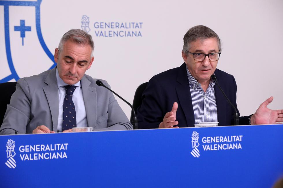 Two men in suits sit at a blue press conference desk with microphones; the man on the left writes while the man on the right speaks, backdrop reads 'Generalitat Valenciana'.