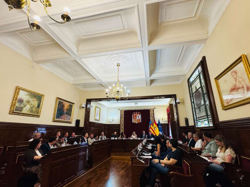 Formal council or board meeting in a wood-paneled chamber with a U-shaped table, flags, and a chandelier overhead.