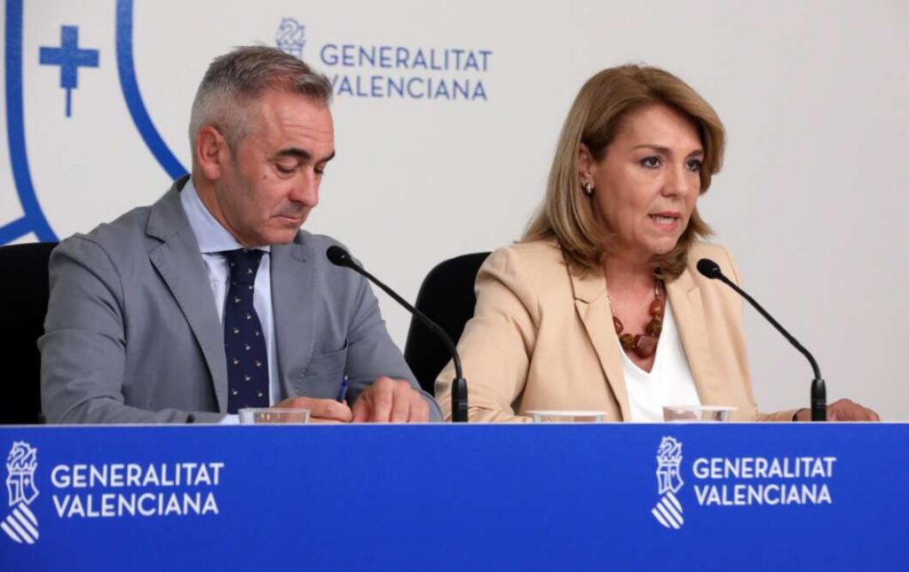 Two officials at a Generalitat Valenciana press conference, seated behind a blue banner with microphones in front of them.