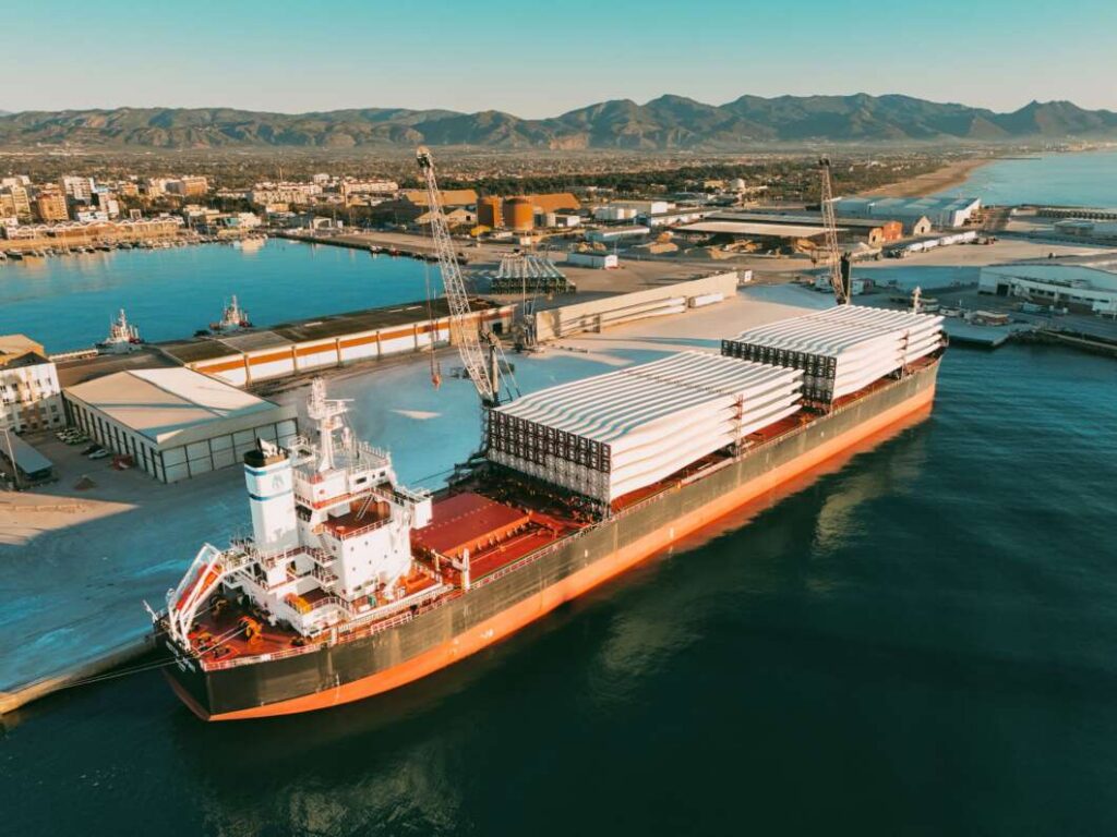 Cargo ship at a port carrying long white wind turbine blades on deck, with cranes and warehouses nearby.