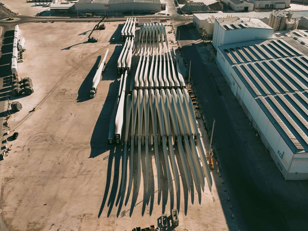 Aerial view of dozens of large wind turbine blades lined up in an open industrial yard beside buildings.