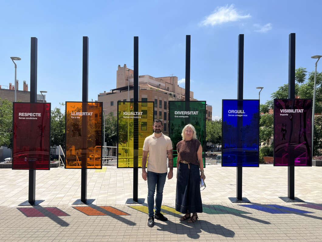 Two people pose in front of a row of tall black posts with colored translucent panels carrying Catalan values (Respecte, Libertat, Igualtat, Diversitat, Orgull, Visibilitat).
