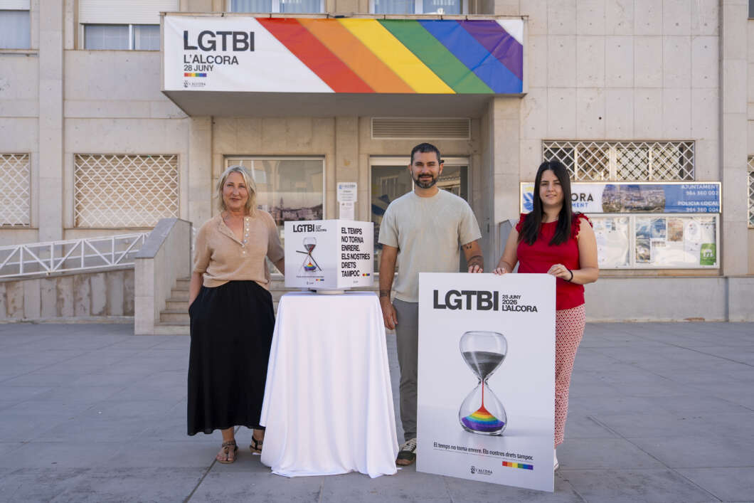 Three people stand outside a building under a rainbow LGTBI banner, posing beside a white-draped table and a large poster with a rainbow-hourglass design on it.