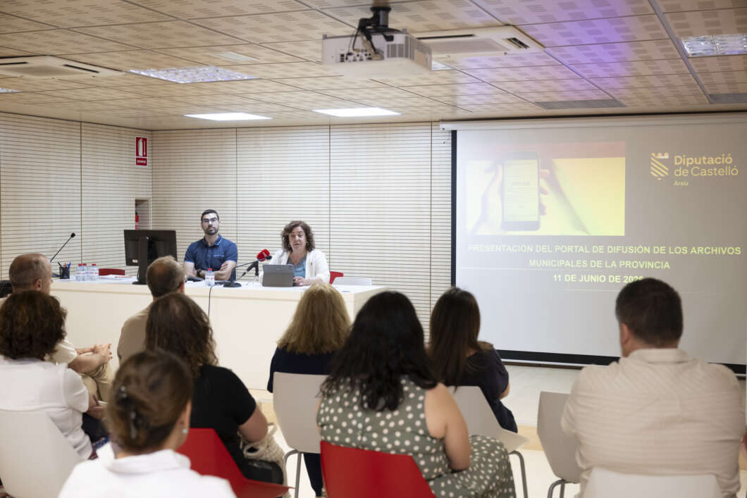 Two presenters sit at a panel desk in a conference room, with a projected slide beside them and an audience watching.